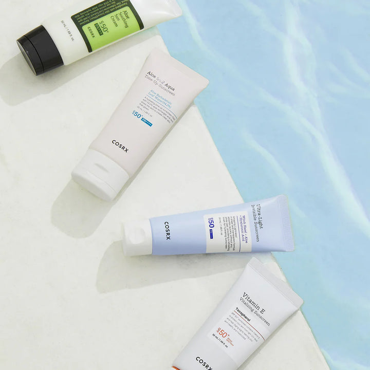 Multiple COSRX sunscreens, including Aloe Soothing Sun Cream, Aloe UV Aqua Tone-Up, Ultra-Light Invisible Sunscreen, and Vitamin E Sunscreen, arranged on a white surface by a pool.