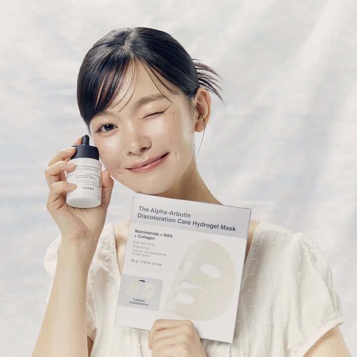 Woman winking while holding COSRX Alpha-Arbutin 2 Discoloration Care Serum and Hydrogel Mask for discoloration and brightening care.