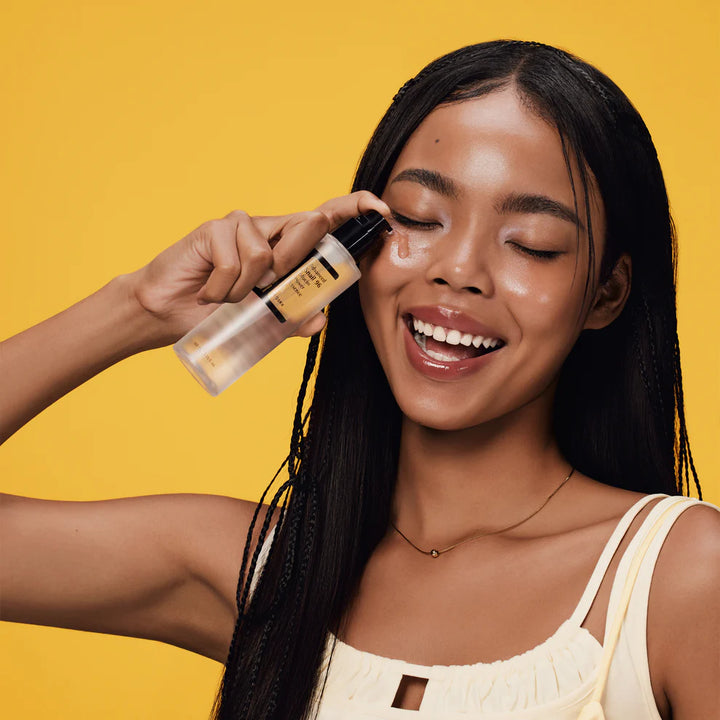 Woman smiling and applying COSRX Advanced Snail 96 Mucin Power Essence on her face against a yellow background.